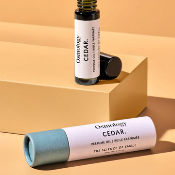 Osmology Cedar perfume oil bottle and its cap displayed on a tan geometric pedestal, with the cylindrical perfume oil tube lying in the foreground against a matching tan background.