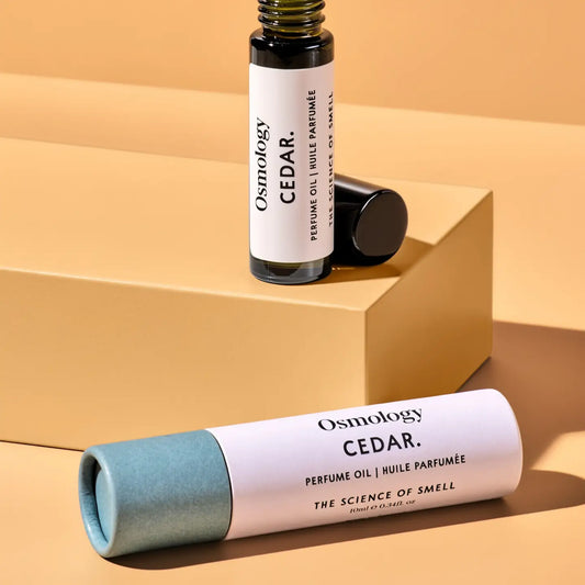 Osmology Cedar perfume oil bottle and its cap displayed on a tan geometric pedestal, with the cylindrical perfume oil tube lying in the foreground against a matching tan background.