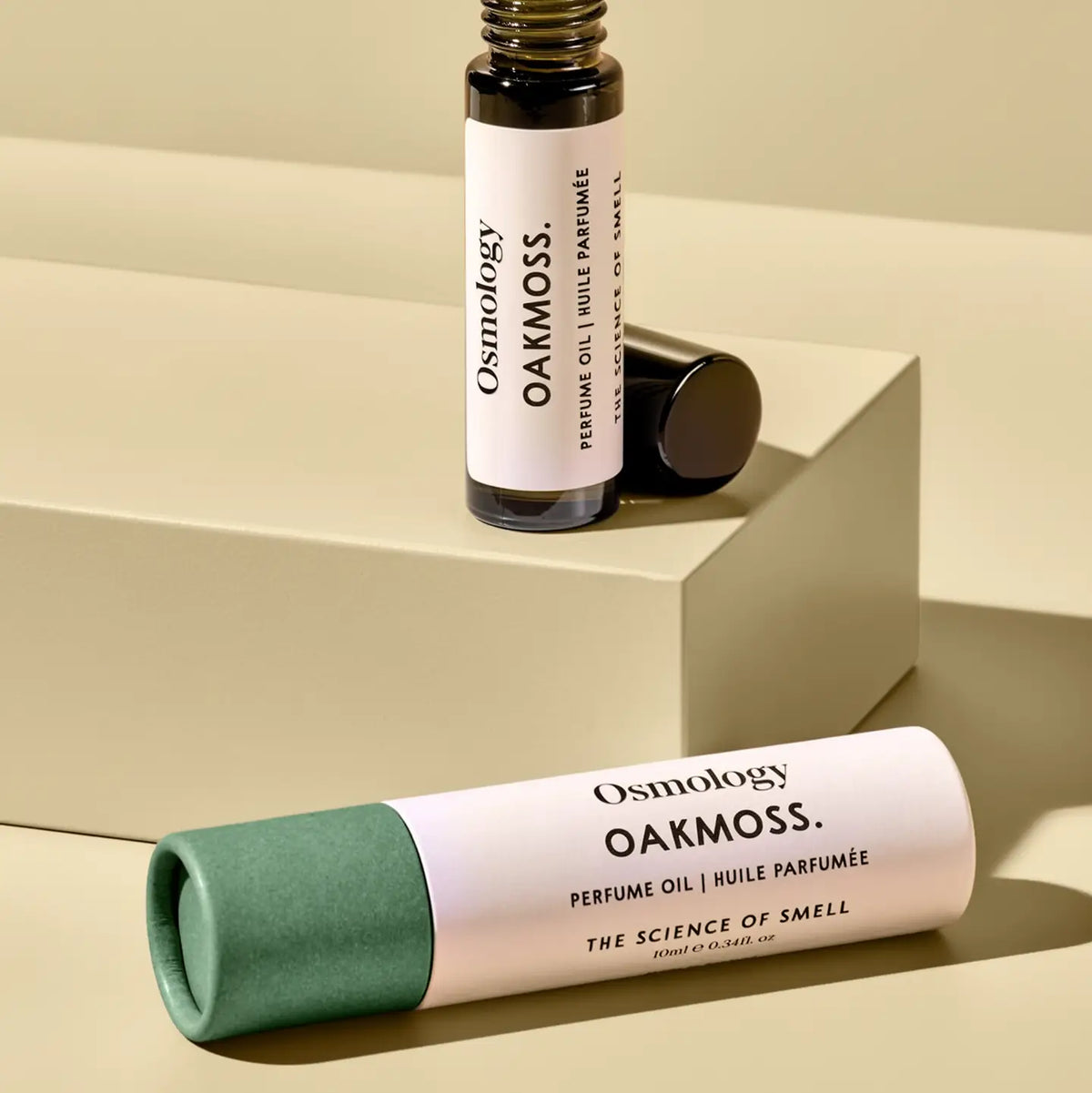 Osmology Oakmoss perfume oil bottle and cap displayed on a beige geometric platform, with a matching cylindrical tube lying in front on a matching beige surface.