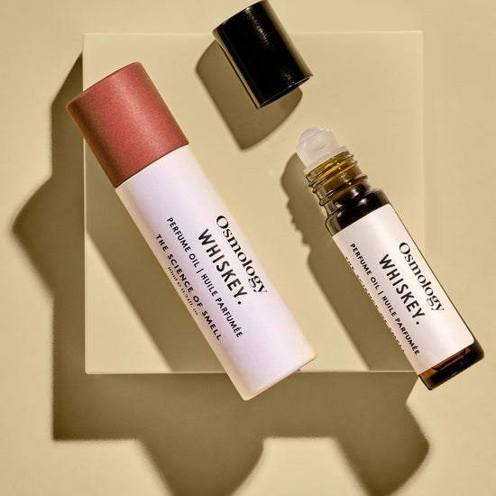 Two cosmetic bottles from Oxology Whiskey, including a white rollerball bottle and a white tube with a pink cap, arranged on a clear square block against a beige background with dramatic shadows.