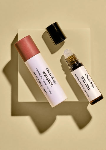Two cosmetic bottles from Oxology Whiskey, including a white rollerball bottle and a white tube with a pink cap, arranged on a clear square block against a beige background with dramatic shadows.