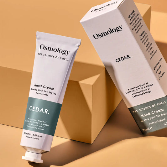 A close-up of a  tube of Osmology Cedar Hand Cream. The tube is grey and teal, with the brand name 'Osmology' and scent 'CEDAR. The box packaging is next to the tube and hey are both balanced on a peach coloured block