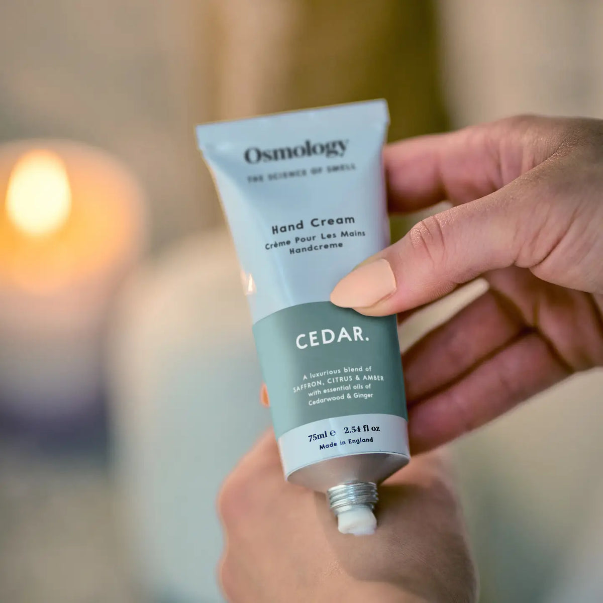 A close-up of a hand holding a squeeze tube of Osmology Cedar Hand Cream. The tube is grey and teal, with the brand name 'Osmology' and scent 'CEDAR.