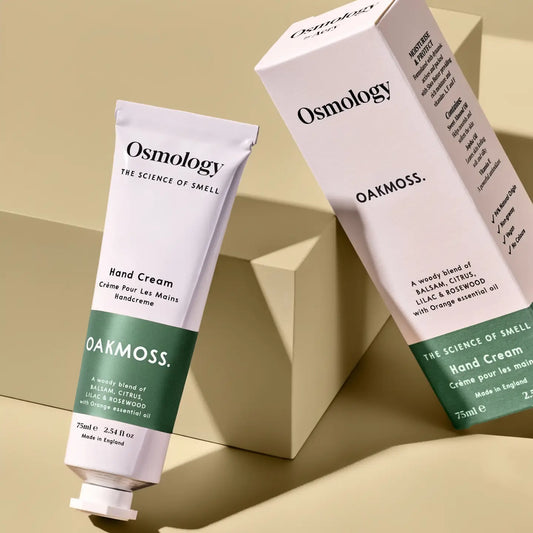 Osmology Oakmoss Hand Cream shown as a white squeeze tube and matching product box on a beige geometric studio background.