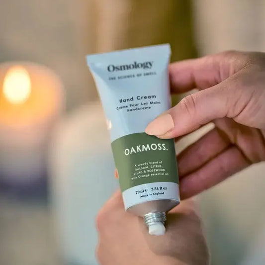 A hand holding an Osmology Oakmoss Hand Cream tube, with a warm, softly blurred background and a glowing candle in the distance.