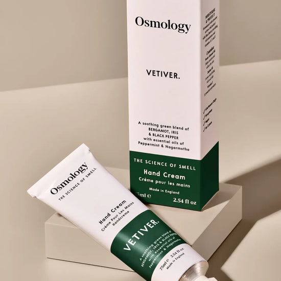 Osmology Vetiver hand cream shown with its white and dark green product box and squeeze tube, arranged on a clean beige surface with a matching pedestal.
