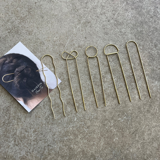 A set of gold-colored brass hairpins laid out on a grey surface, next to a business card with a how to use guide 