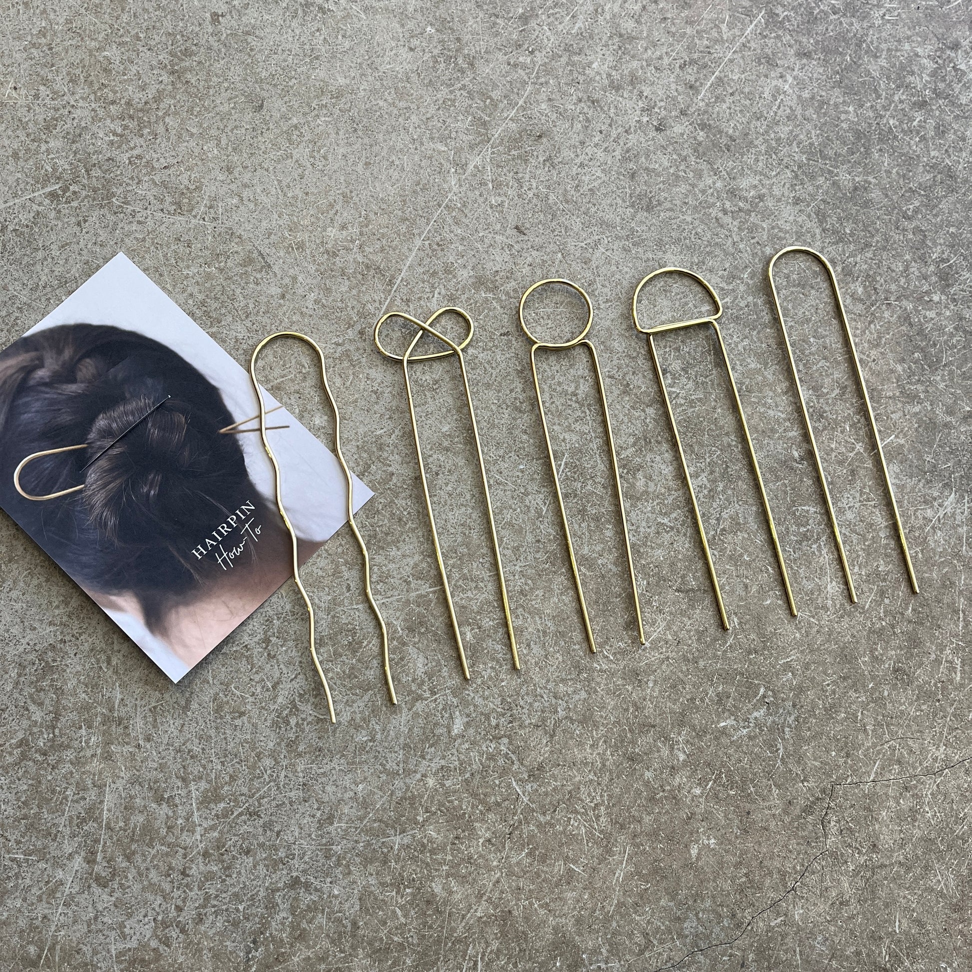 A set of gold-colored brass hairpins laid out on a grey surface, next to a business card with a how to use guide 