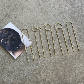 A set of gold-colored brass hairpins laid out on a grey surface, next to a business card with a how to use guide 