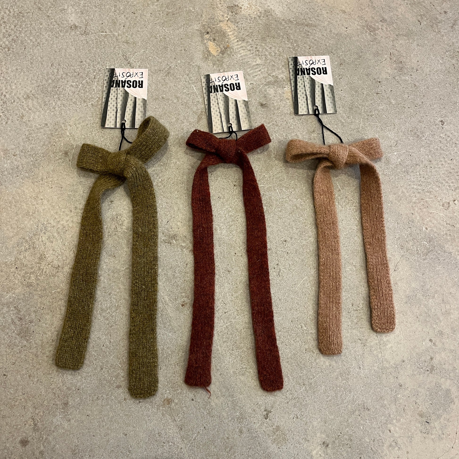 Three knitted bows in green, red, and brown on a concrete surface with labels.