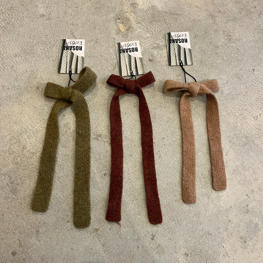 Three knitted bows in green, red, and brown on a concrete surface with labels.