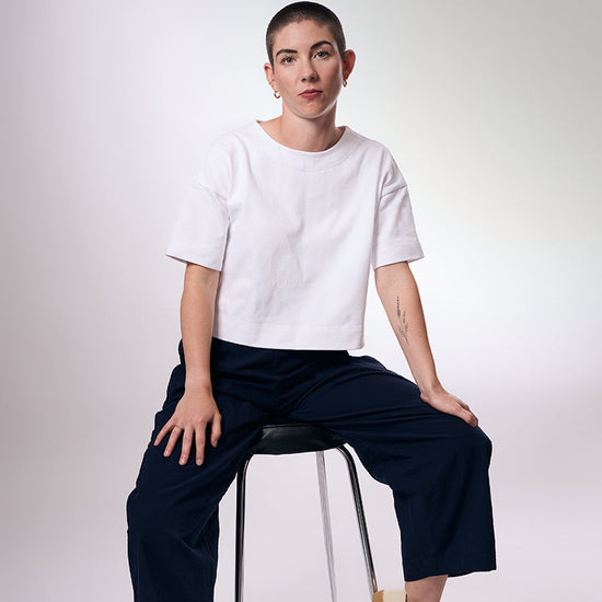 Person sitting on a chair wearing a white t-shirt and navy pants against a plain background