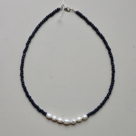 A beaded necklace featuring a strand of tiny dark beads with a row of five white freshwater pearls at the bottom, finished with a silver clasp, shown on a neutral light gray background.