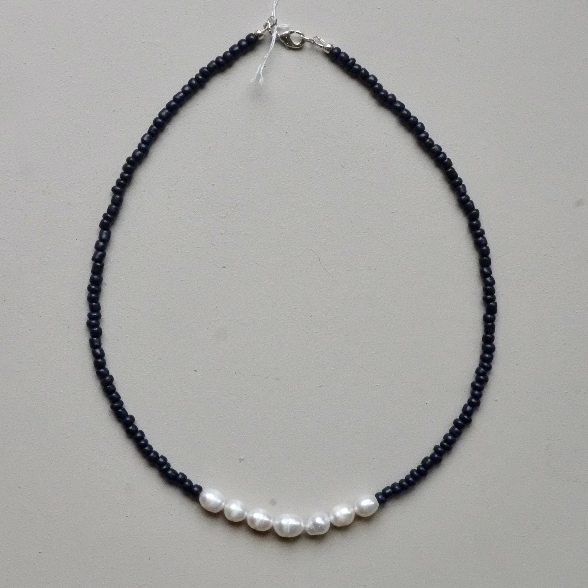 A beaded necklace featuring a strand of tiny dark beads with a row of five white freshwater pearls at the bottom, finished with a silver clasp, shown on a neutral light gray background.