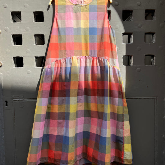 Brightly coloured sleeveless checked dress with gathered waist and round neck hanging against a medieval grey door on a hanger 