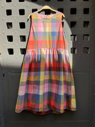 Brightly coloured sleeveless checked dress with gathered waist and round neck hanging against a medieval grey door on a hanger 