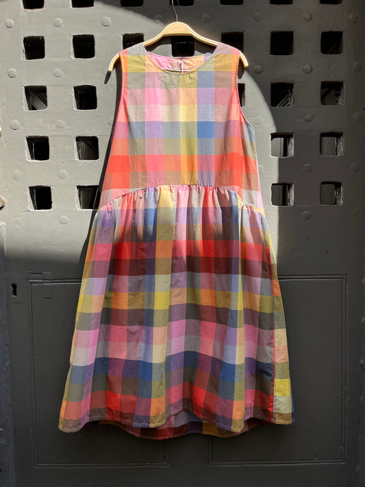 Brightly coloured sleeveless checked dress with gathered waist and round neck hanging against a medieval grey door on a hanger 
