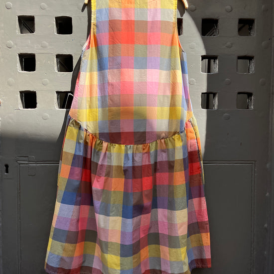 A sleeveless tiered dress with a colorful plaid pattern, hanging on a wooden hanger against a dark grey wooden door with square cutouts.