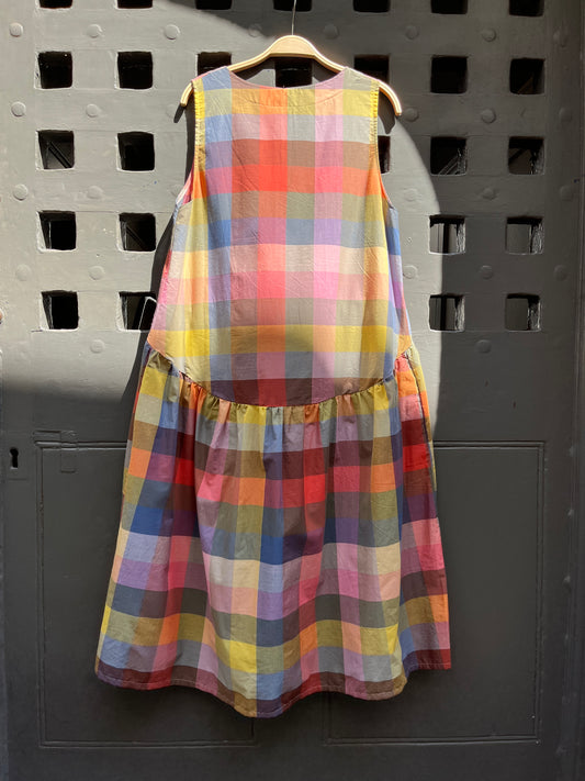 A sleeveless tiered dress with a colorful plaid pattern, hanging on a wooden hanger against a dark grey wooden door with square cutouts.