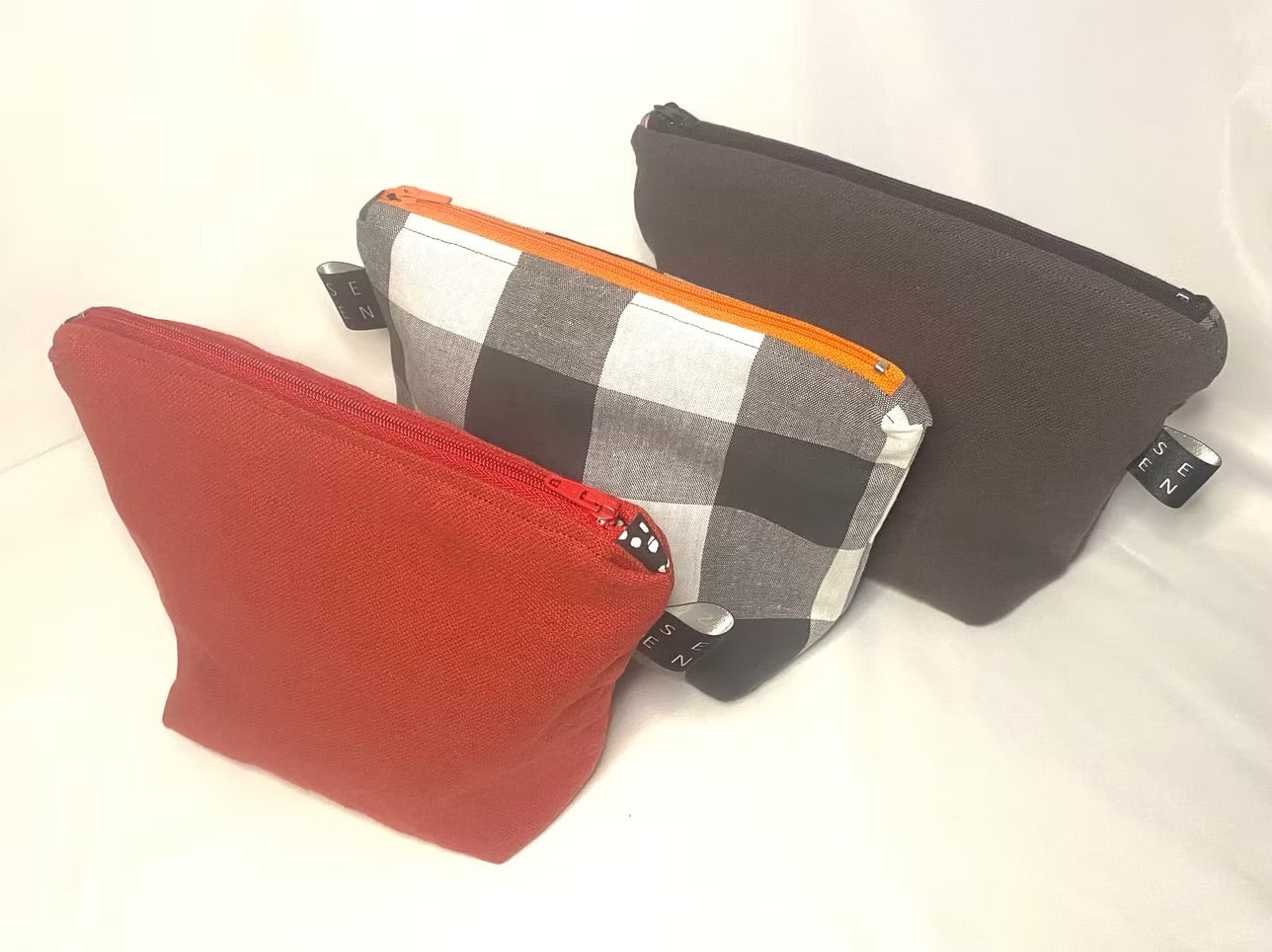 Three pouches in red, black and white checkered, and black with orange zipper on a white background 