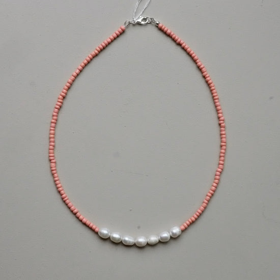 A delicate beaded necklace featuring a strand of small pinkish-orange beads with five white baroque pearls aligned across the front, finished with a silver clasp, shown on a neutral gray background.