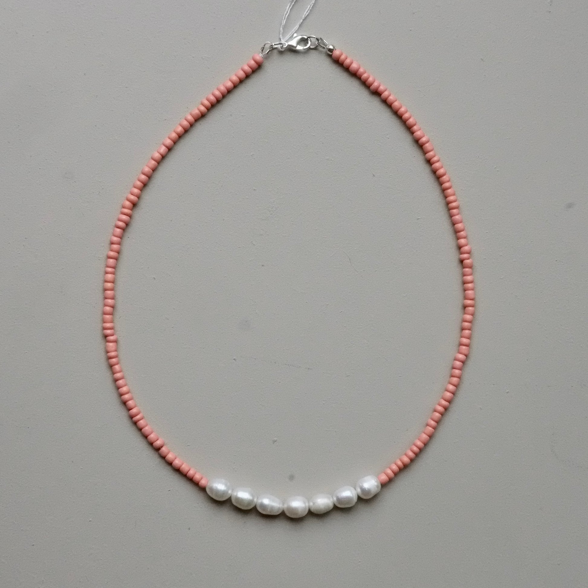 A delicate beaded necklace featuring a strand of small pinkish-orange beads with five white baroque pearls aligned across the front, finished with a silver clasp, shown on a neutral gray background.