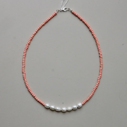 A delicate beaded necklace featuring a strand of small pinkish-orange beads with five white baroque pearls aligned across the front, finished with a silver clasp, shown on a neutral gray background.