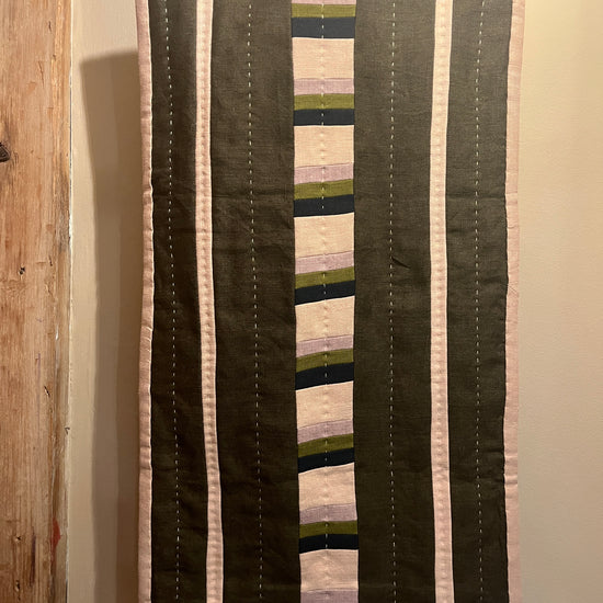 Khaki, mauve, navy and beige stripe quilted wall hanging with white stitch detail hanging on a beige wall