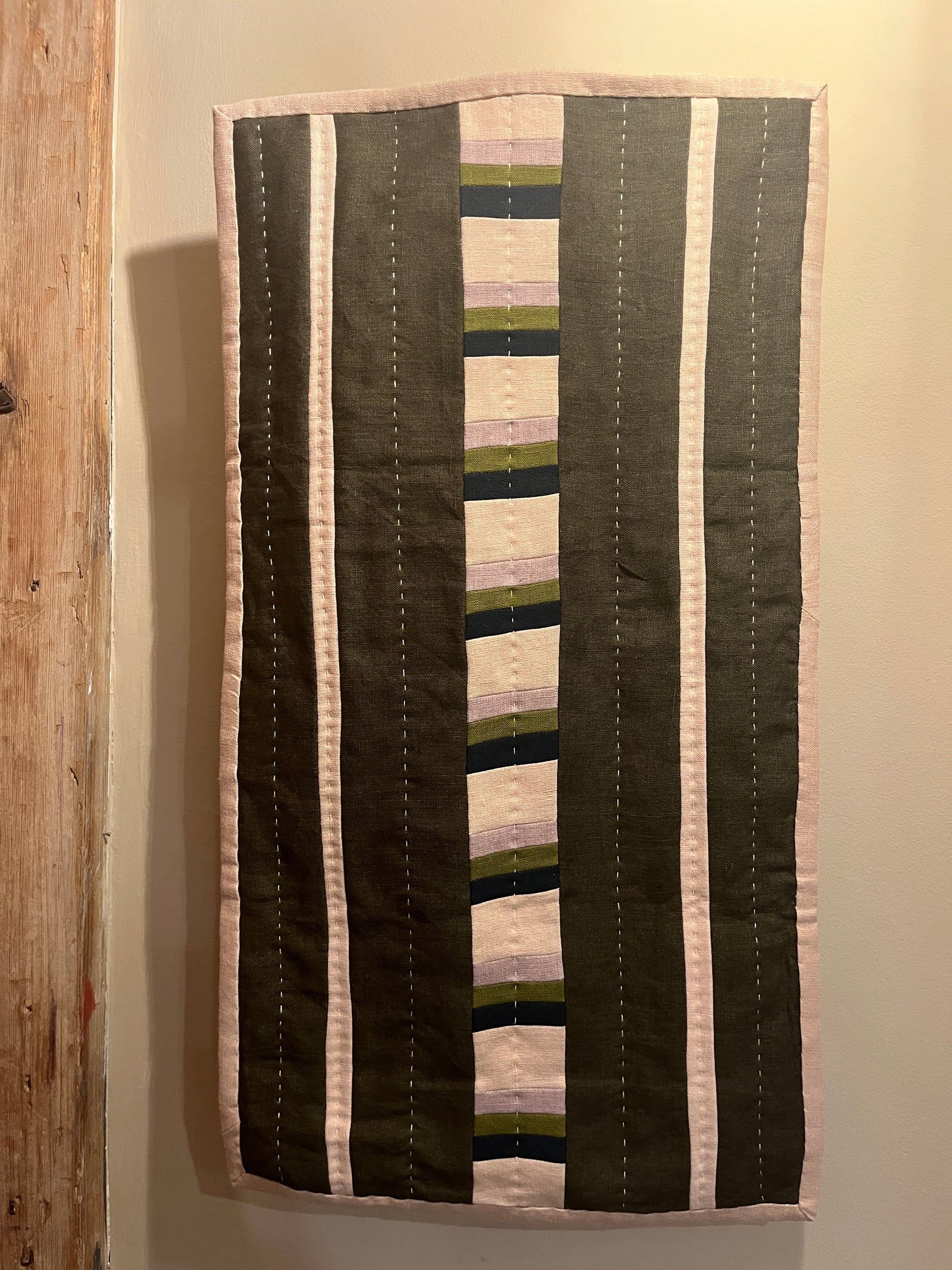 Khaki, mauve, navy and beige stripe quilted wall hanging with white stitch detail hanging on a beige wall