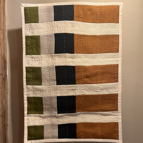 A handmade patchwork quilt hanging on a neutral beige wall. The quilt features a grid pattern with vertical stripes in orange, cream, black, and light gray, and horizontal stripes in olive green and cream.