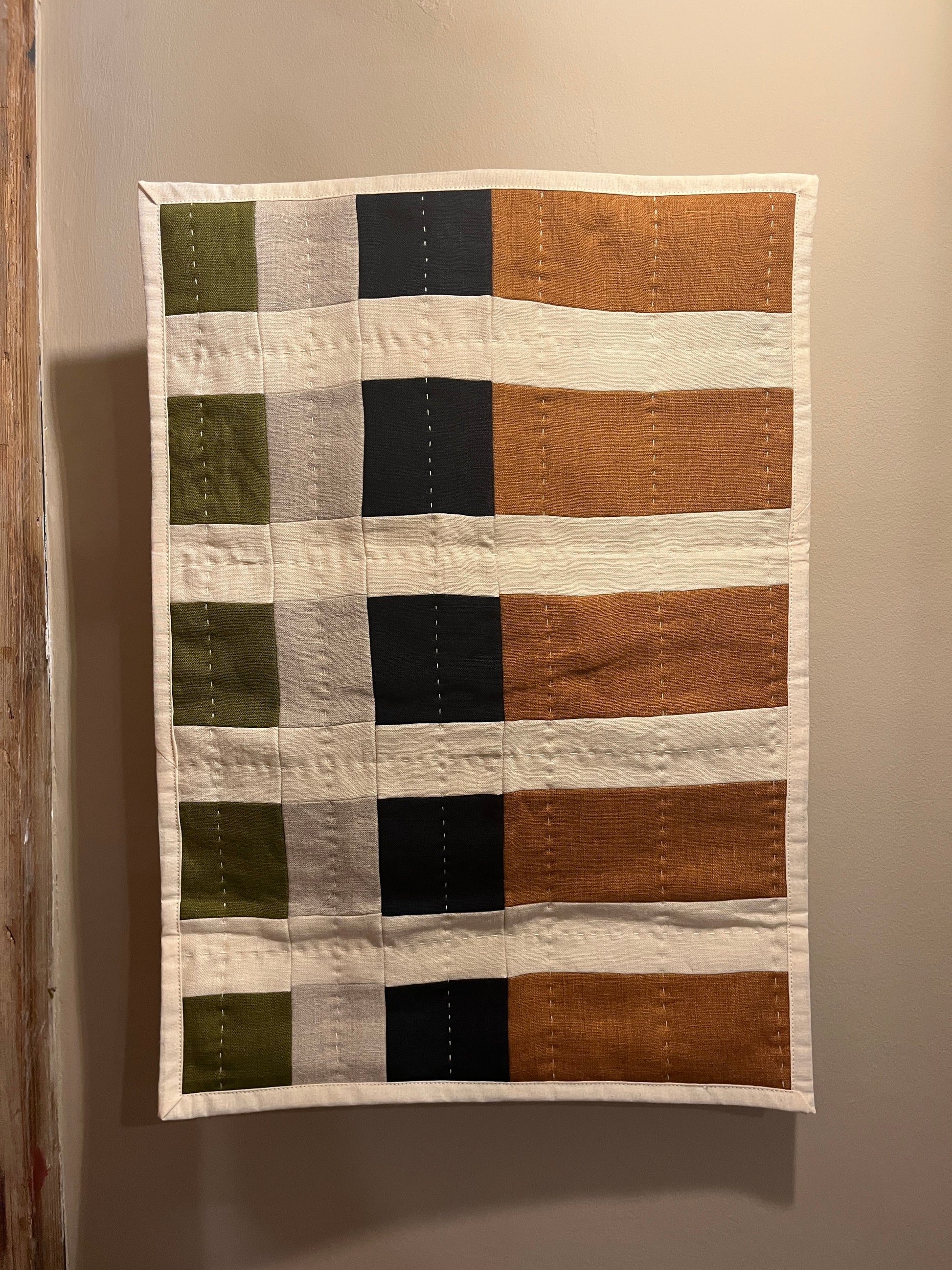 A handmade patchwork quilt hanging on a neutral beige wall. The quilt features a grid pattern with vertical stripes in orange, cream, black, and light gray, and horizontal stripes in olive green and cream.