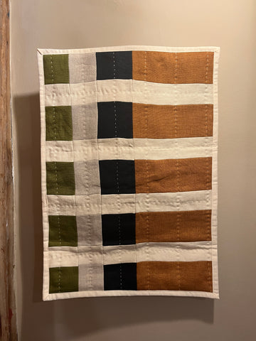 A handmade patchwork quilt hanging on a neutral beige wall. The quilt features a grid pattern with vertical stripes in orange, cream, black, and light gray, and horizontal stripes in olive green and cream.