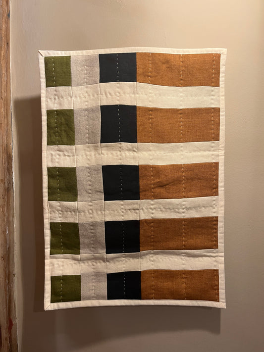 A handmade patchwork quilt hanging on a neutral beige wall. The quilt features a grid pattern with vertical stripes in orange, cream, black, and light gray, and horizontal stripes in olive green and cream.