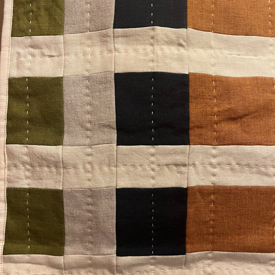 A close-up of a patchwork quilted fabric with a grid pattern of vertical and horizontal stripes in orange, black, light gray, olive green, and cream.