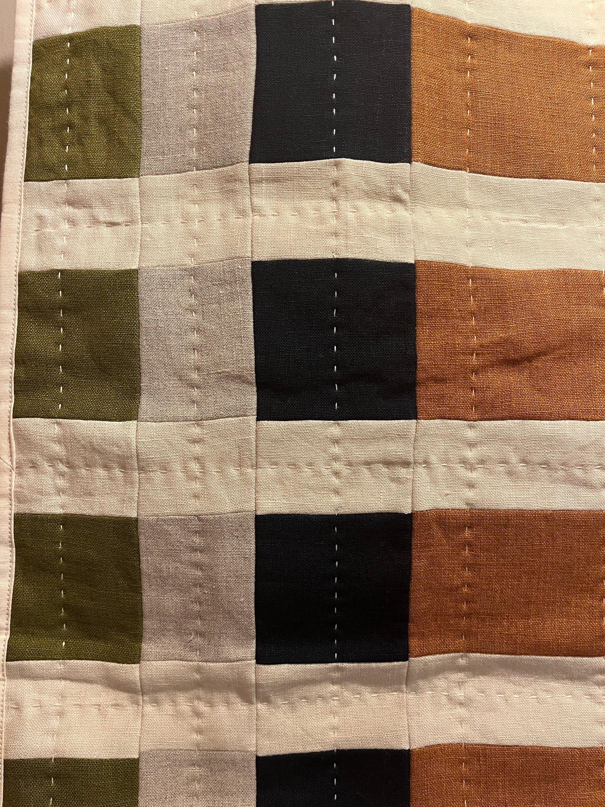 A close-up of a patchwork quilted fabric with a grid pattern of vertical and horizontal stripes in orange, black, light gray, olive green, and cream.