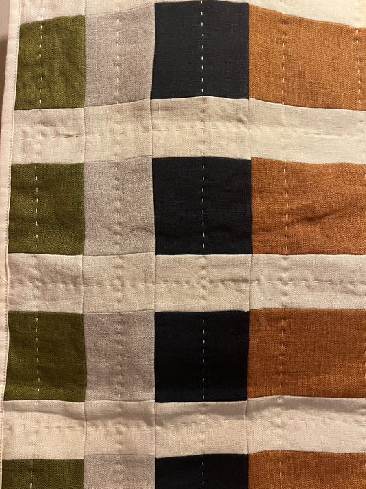 A close-up of a patchwork quilted fabric with a grid pattern of vertical and horizontal stripes in orange, black, light gray, olive green, and cream.