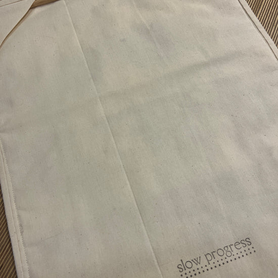 A close-up of a light beige fabric all hanging back featuring a diagonal fold and the printed brand name 'slow process' in black text with a dotted design in the upper right corner.