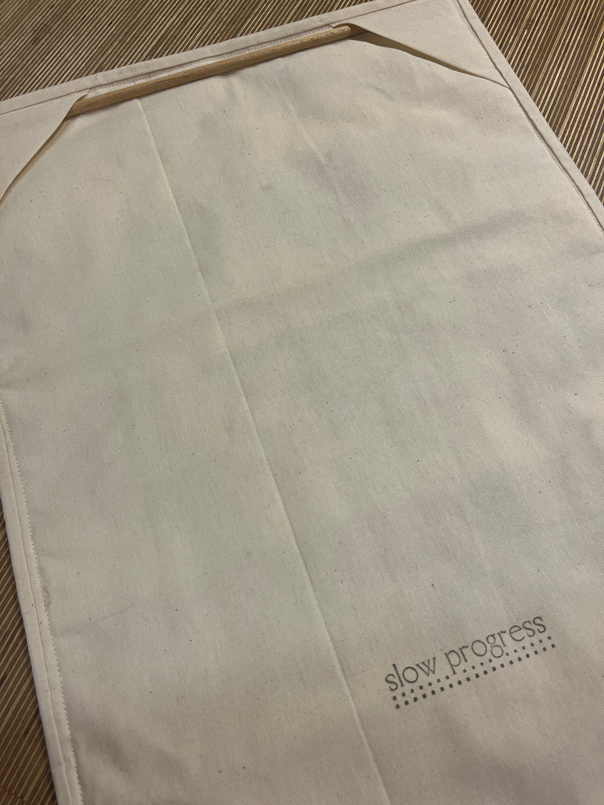 A close-up of a light beige fabric all hanging back featuring a diagonal fold and the printed brand name 'slow process' in black text with a dotted design in the upper right corner.