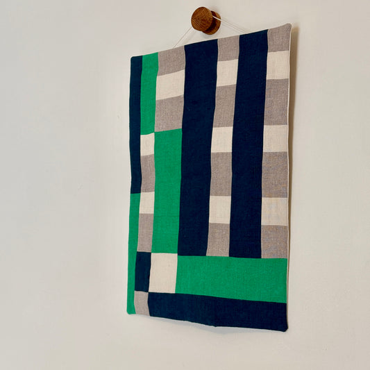 Checkered fabric wall hanging with green, blue, and gray pattern on a plain background
