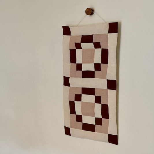 Patchwork wall hanging with brown and beige squares on a plain wall.