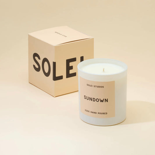 A white candle labeled 'Sundown' by Solei Studios, next to its packaging box with the word 'SOLEI' on it, all set against a beige background.