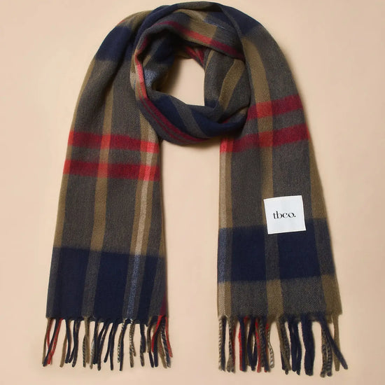 A tartan scarf with navy blue, beige, brown, and red stripes, featuring fringed ends and a white brand label that reads 'tbco.' on a solid beige background.