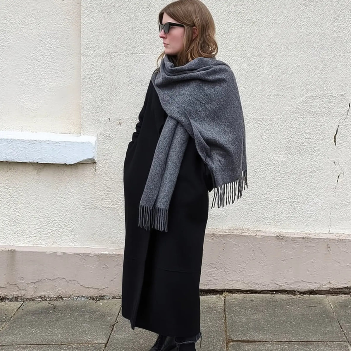 A woman wearing a large grey knitted scarf with fringe over a long black coat, standing outdoors against a textured off-white wall.