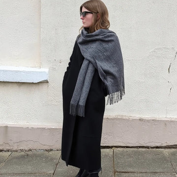 A woman wearing a large grey knitted scarf with fringe over a long black coat, standing outdoors against a textured off-white wall.