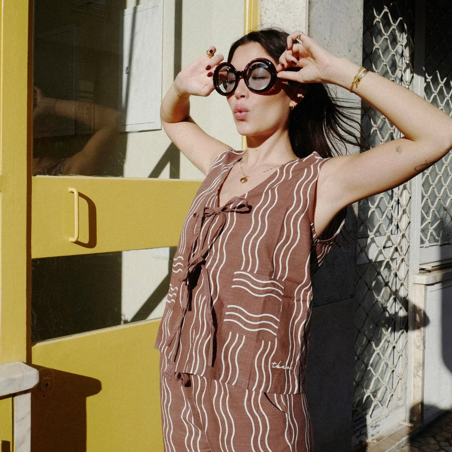 Woman in a patterned wavy brown and white top and trousers standing in front of a yellow door.