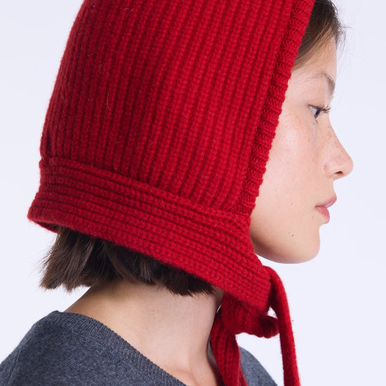A side-profile portrait of a person wearing a bright red ribbed knit bonnet with ties, against a clean light gray background.