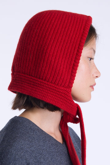 A side-profile portrait of a person wearing a bright red ribbed knit bonnet with ties, against a clean light gray background.