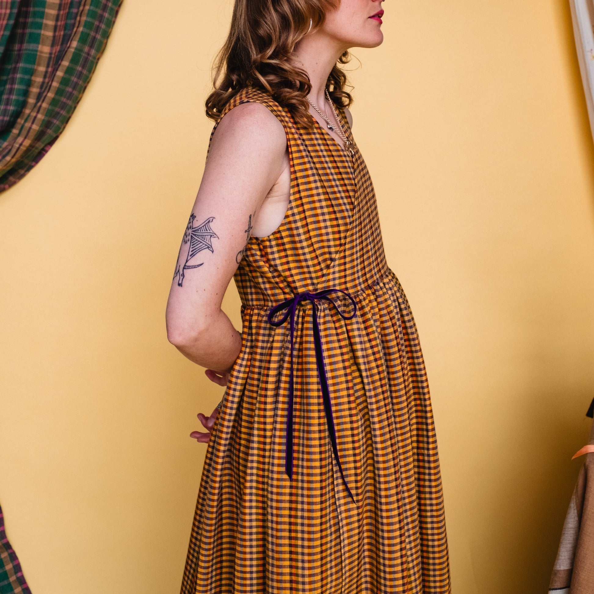 Woman in a checkered dress standing against a yellow wall with a plaid curtain.
