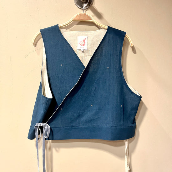 A sleeveless wrap-style vest in indigo blue fabric with white ties and white lining, hanging on a wooden hanger against a beige wall.