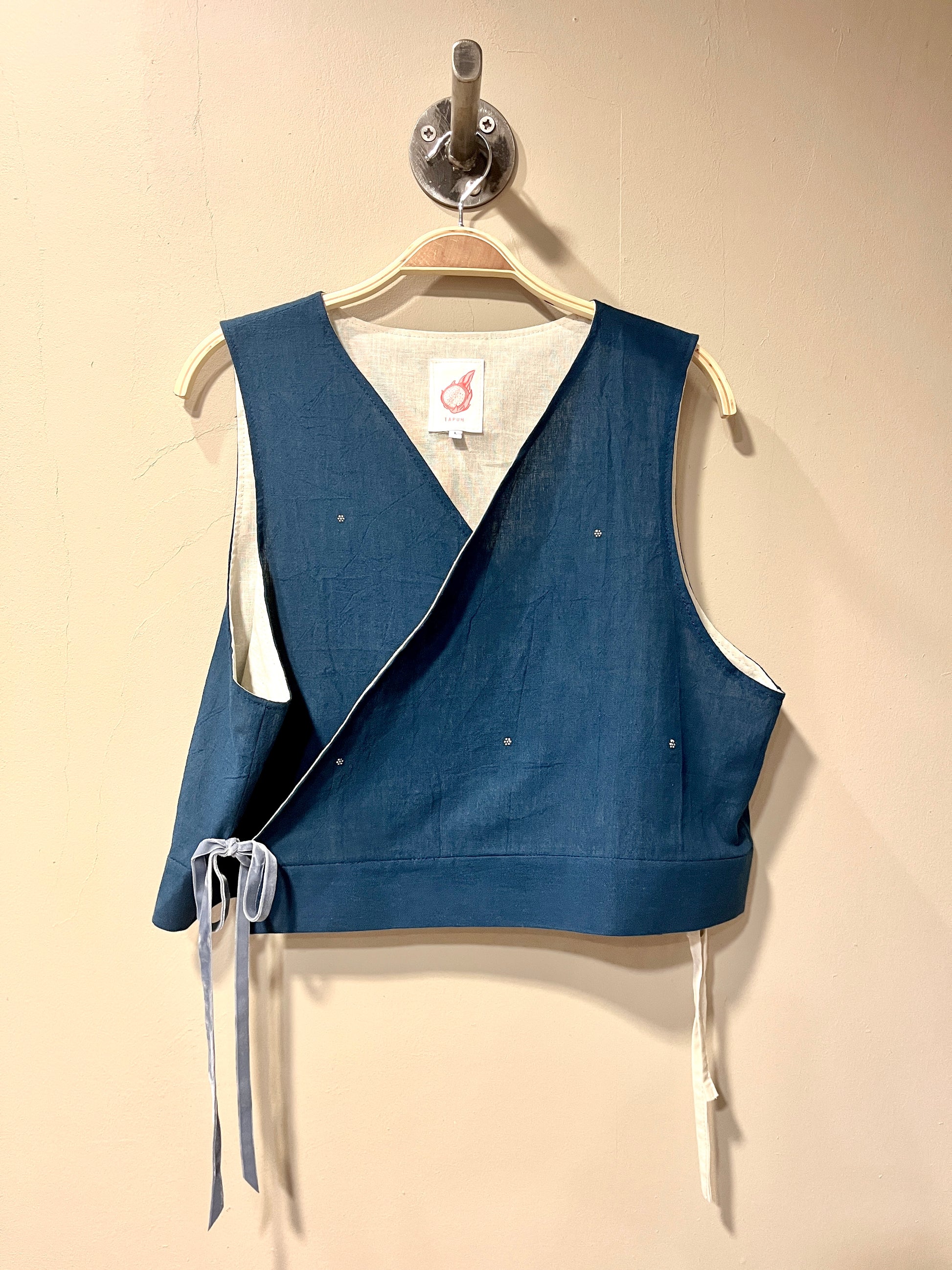 A sleeveless wrap-style vest in indigo blue fabric with white ties and white lining, hanging on a wooden hanger against a beige wall.